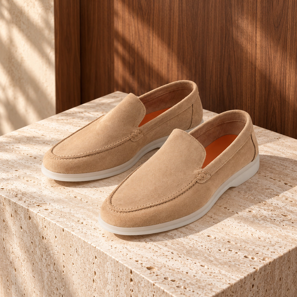 Loafers cappuccino din piele întoarsă bej, cu talpă albă, așezați pe un soclu de travertin.